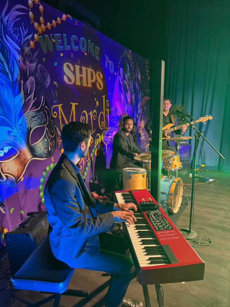 Trio on stage with red keys, drums, and electric bass in front of a purple “Welcome to SHPS Mardi Gras” backdrop and green drapes