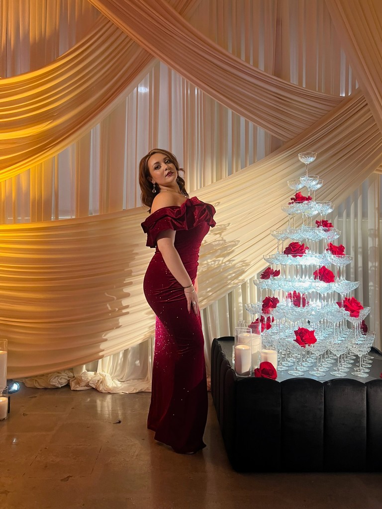 Stephanie Salahz in a burgundy velvet gown beside a glowing champagne coupe tower with roses and candles