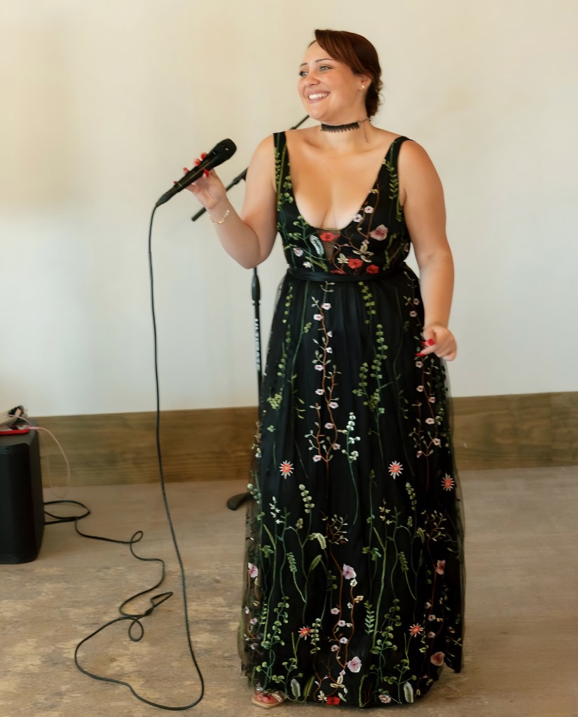 Stephanie Salahz in a floor-length black gown with floral embroidery, holding a microphone and smiling