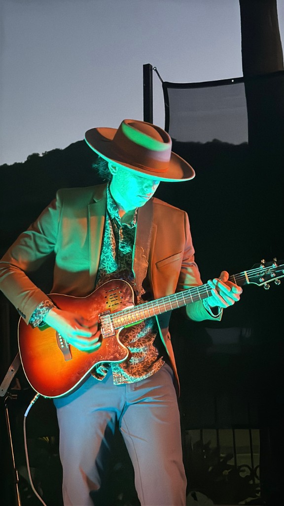 Guitarist in a wide-brimmed hat playing a sunburst semi-hollow guitar under vivid green stage light at dusk, hills silhouetted behind him