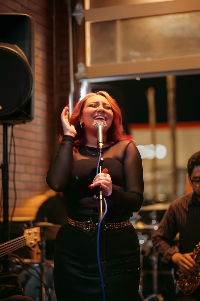 Stephanie Salahz in a black lace-sleeve outfit singing at a microphone with a saxophonist beside her
