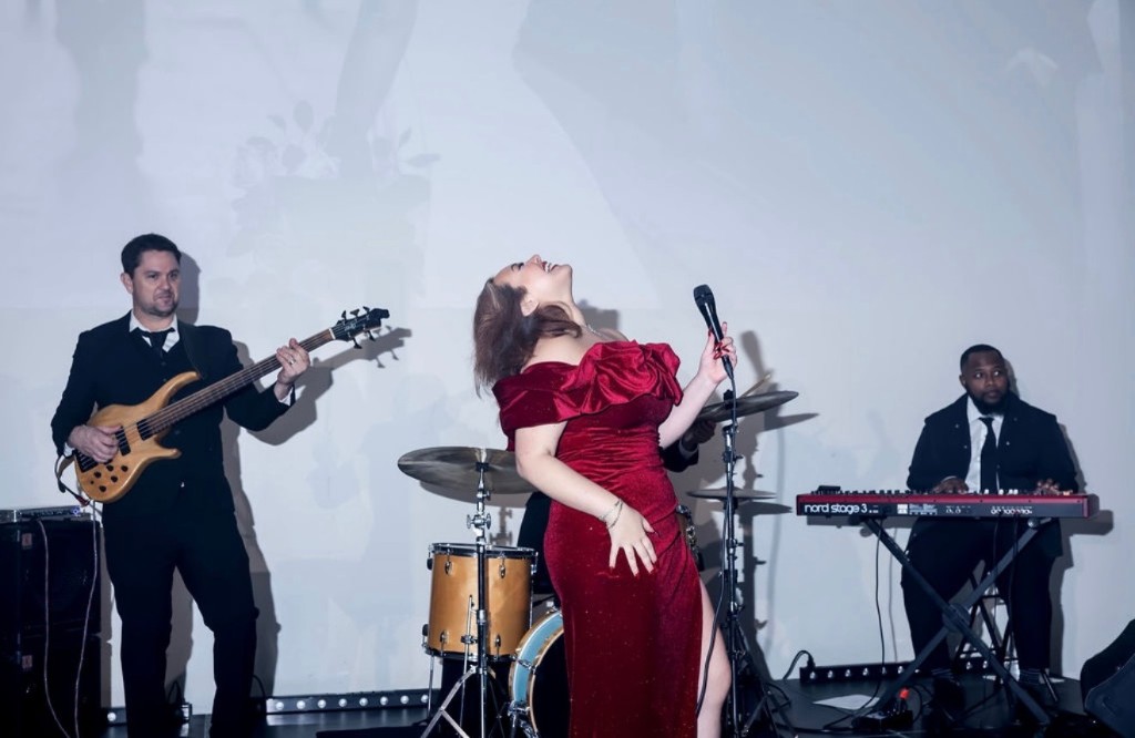 Stephanie Salahz in a red velvet gown singing between bassist and keyboardist on a bright lit stage