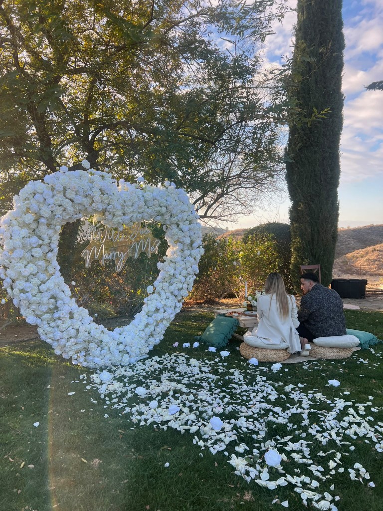 Outdoor garden scene with a white rose heart arch and neon “Will You Marry Me?” sign, couple seated at a picnic table