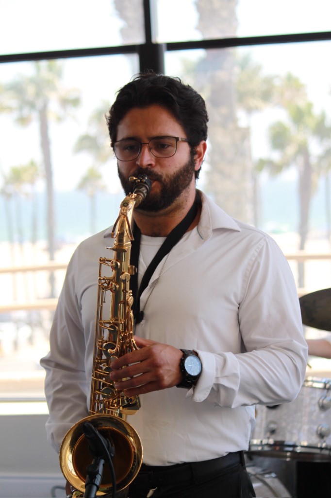 Saxophonist in a white shirt playing a gold saxophone indoors, bright beach and palm trees visible through a large window behind him