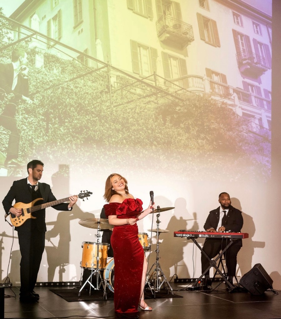 Stephanie Salahz in a red velvet gown singing on stage with bass and Nord keys, soft building projection behind