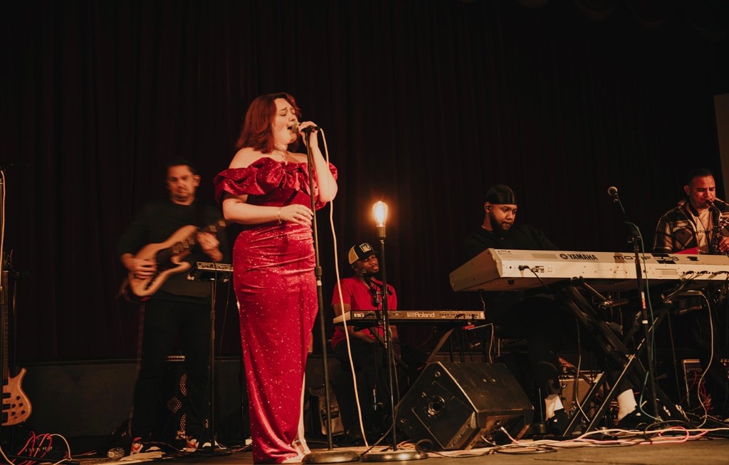 Stephanie Salahz singing on stage in a red gown with bass, keyboards, and saxophone
